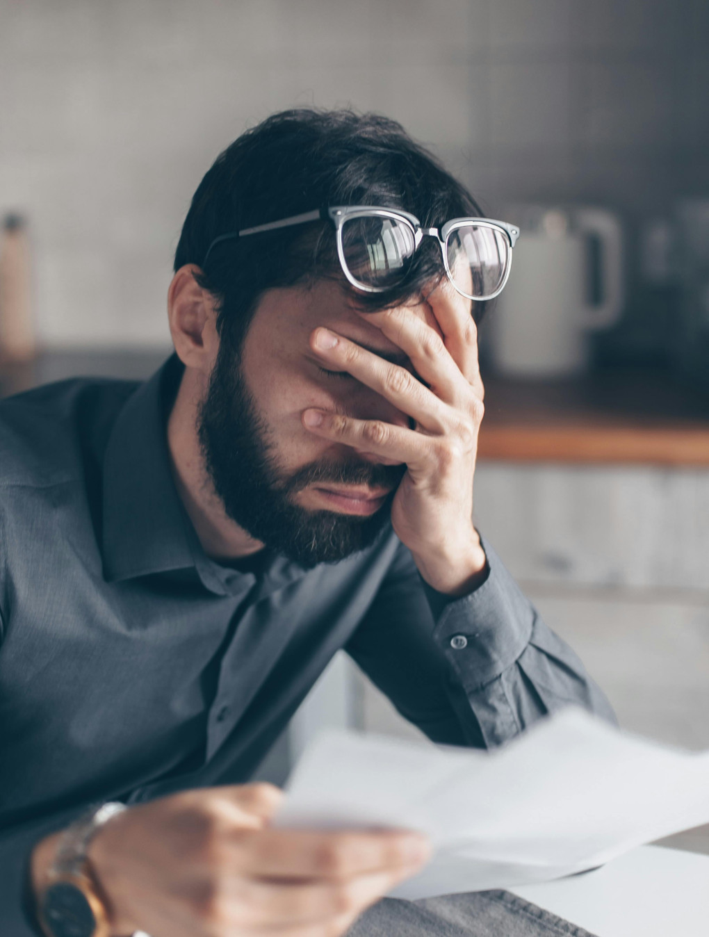 Das Bild zeigt einen Mann mit dunklem Haar und Bart, der eine Brille auf dem Kopf trägt. Er sitzt an einem Tisch und hält ein Blatt Papier in der Hand. Mit der anderen Hand bedeckt er sein Gesicht, es wirkt so als ob er gestresst oder frustriert ist. Im Hintergrund ist eine verschwommene Küche zu sehen, einschließlich eines Wasserkochers